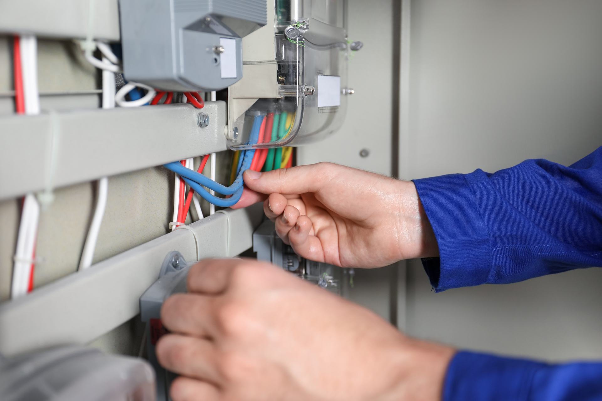 Hands in blue work sleeves connecting coloured wires inside a grey electrical enclosure